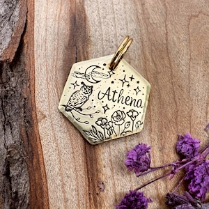 Puede incluir: Un colgante hexagonal de color crema con un anillo dorado. El colgante presenta una ilustración negra de un búho, luna, estrellas, flores y el nombre "Athena". El colgante se muestra sobre una superficie de madera con flores moradas.