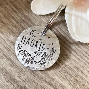 May include: Silver key chain with a round tag featuring a moon, stars, mountains, trees, and the name "HAGRID" engraved on it.