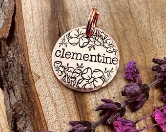 May include: A round, copper-colored pet tag with the name "clementine" in a script font, surrounded by a floral design. The tag has a copper-colored ring for attachment. The background is a wooden surface with dried purple flowers.