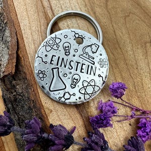Puede incluir: Una chapa redonda de metal plateado para llavero con el nombre "EINSTEIN" en letras negras. La chapa está decorada con ilustraciones de temática científica, incluyendo átomos, bombillas y vasos de precipitados. Un llavero plateado está adjunto. El fondo es una superficie de madera con flores moradas.