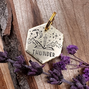 May include: A brass dog tag with a hexagonal shape, featuring a black and white engraved design of a dragon, a sword, a book, and mountains. The tag has the word "THUNDER" engraved on the bottom.
