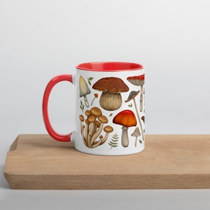 Puede incluir: Taza de cerámica blanca con borde rojo, con una ilustración colorida de varios hongos. La taza está sobre una superficie de madera.
