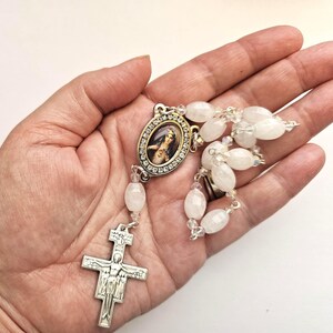 May include: A rosary featuring translucent white oval beads and silver-toned metal links. The centrepiece is an oval pendant with a portrait of the Sacred Heart of Jesus, framed by small clear rhinestones. A silver-toned crucifix hangs at the end.