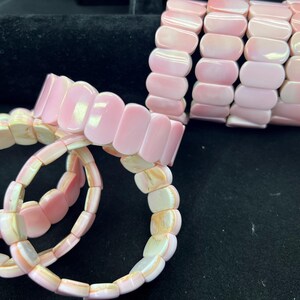 Op de afbeelding: Verschillende roze en witte kralenarmbanden worden gepresenteerd tegen een zwarte achtergrond. De armbanden hebben een rechthoekig kralenontwerp. De kleuren zijn zacht en vrouwelijk, met een parelmoerglans. De armbanden zijn waarschijnlijk mode- of sieradenaccessoires.