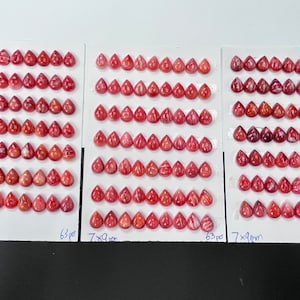May include: A collection of 63 teardrop-shaped cabochons, each measuring 7x9 mm, arranged in rows on a white card. The cabochons are a vibrant red color with white streaks throughout.