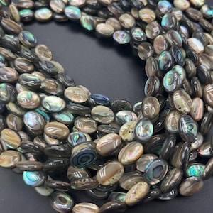 May include: A collection of oval-shaped abalone shell beads featuring a smooth, polished surface with iridescent swirls of dark brown, grey, and vibrant green and blue tones. The beads are arranged in a dense, curved cluster, showcasing their natural, multi-coloured lustre and unique, organic patterns on each individual piece.