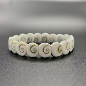 Puede incluir: Un brazalete blanco de conchas con un patrón en espiral. El brazalete está hecho de múltiples conchas pequeñas y redondas que están enhebradas.