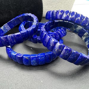 以下が含まれることがあります： 青いラピスラズリジェムストーンのブレスレットが4つ、重ねられています。ブレスレットは正方形のビーズで作られています。