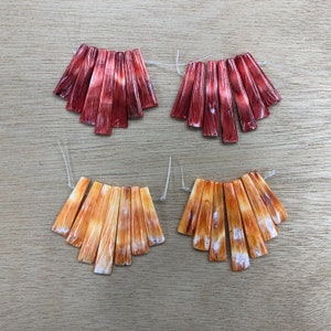 May include: Two sets of eight shell pieces, each set in a different color. One set is a reddish-brown color and the other set is a light orange color. The shell pieces are arranged in a fan shape.