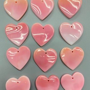 May include: Twelve pink heart-shaped shell pendants with a hole at the top for hanging. The shells have a pearlescent finish and are arranged in three rows of four.