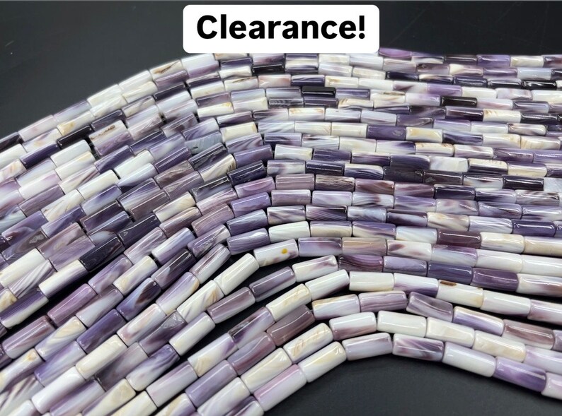 May include: A strand of purple and white shell beads. The beads are rectangular and have a mottled pattern. The beads are arranged in a curved line.