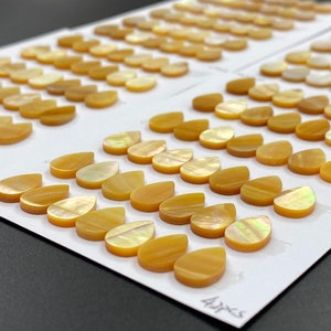 May include: A close-up of a white surface with rows of iridescent, teardrop-shaped, mother-of-pearl guitar inlays. The inlays are arranged in rows and are labeled with the measurements "8x12mm" and "4x7.5".