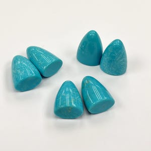 May include: Six turquoise-colored, cone-shaped stones are arranged on a white surface. The stones have a smooth, polished appearance with subtle veining. The rounded bases and pointed tops give them a unique, organic shape.