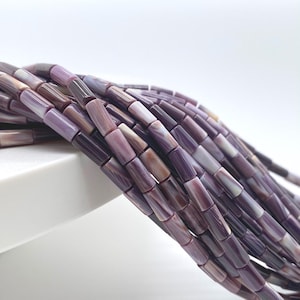 May include: A collection of cylindrical beads in varying shades of purple, lavender, and deep plum. Each bead features a smooth, polished surface with subtle, natural-looking striations and a glossy finish, arranged in multiple parallel strands.
