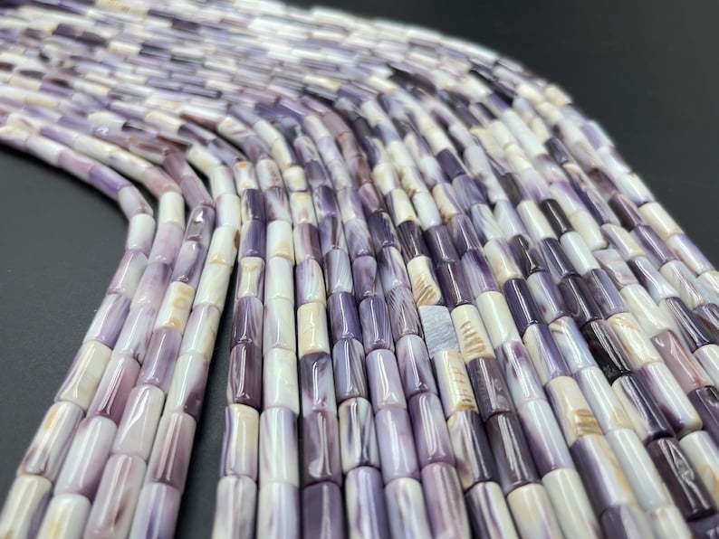 May include: A strand of purple and white shell beads, each bead is a cylinder shape with a smooth, polished finish.