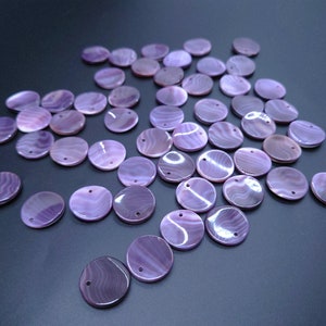 May include: A collection of purple shell round beads with a hole in the center. The beads are scattered on a dark gray surface.