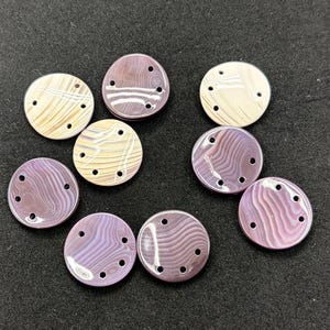 May include: A collection of nine circular buttons featuring a smooth, polished finish with swirling patterns in shades of purple, lavender, and cream. Each button is designed with four small holes arranged in a curved line for attachment.