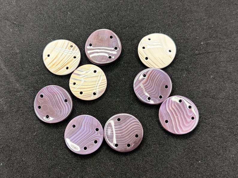 May include: A collection of nine circular buttons featuring a smooth, polished finish with swirling patterns in shades of purple, lavender, and cream. Each button is designed with four small holes arranged in a curved line for attachment.
