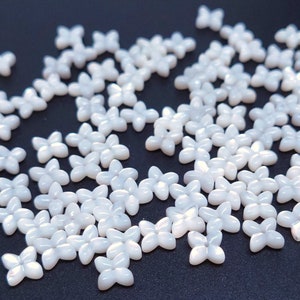 Puede incluir: Un conjunto de pequeñas cuentas blancas con forma de flor de cuatro pétalos dispersas sobre una superficie oscura. Las cuentas tienen un brillo nacarado, que refleja la luz. Estas cuentas decorativas son ideales para manualidades y bisutería.