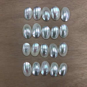 Peut inclure: Une collection de 18 cabochons ovales en nacre blanche irisée. Les cabochons sont disposés en trois rangées de six, chaque rangée étant légèrement décalée par rapport à celle du dessus.