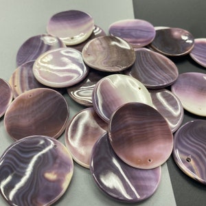 May include: A collection of round, flat shell pendants featuring a smooth, polished surface with swirling patterns in shades of purple, lavender, and white. Each circular piece has a small, pre-drilled hole near the edge for threading or attachment.