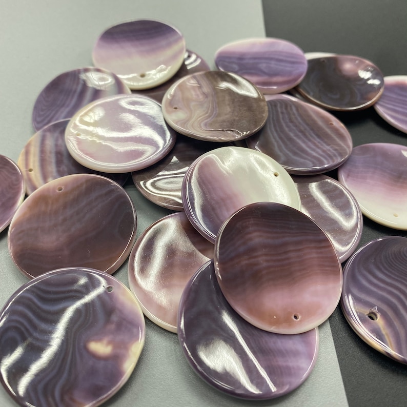 May include: A collection of round, flat shell pendants featuring a smooth, polished surface with swirling patterns in shades of purple, lavender, and white. Each circular piece has a small, pre-drilled hole near the edge for threading or attachment.