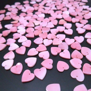 May include: A collection of small, heart-shaped beads in varying shades of pink. Each bead has a small hole at the top, suggesting they are designed for stringing. The beads are scattered on a dark surface, creating a visually appealing pattern.