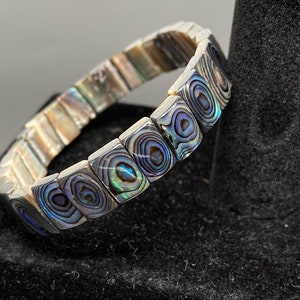 Könnte beinhalten: Ein Armband aus schillernden Abalone-Muschelplatten. Die Platten sind rechteckig und haben ein wirbelndes Muster. Das Armband liegt auf einer schwarzen Oberfläche.