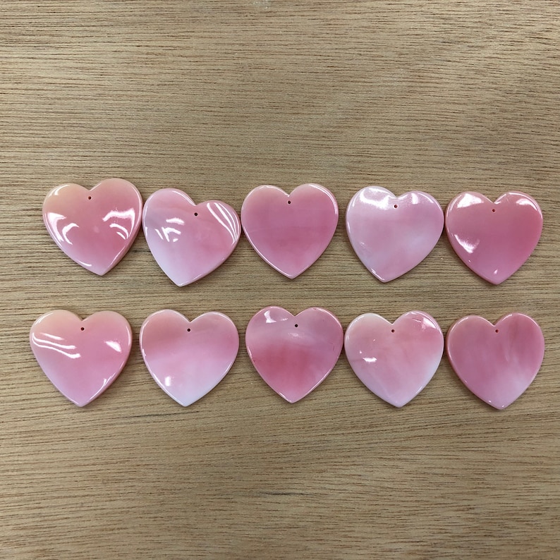 May include: Ten pink heart-shaped shell charms with a hole at the top for stringing. The charms are made of natural shell and have a glossy finish.