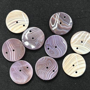 May include: A collection of eight round buttons featuring a smooth, glossy finish with swirling patterns in shades of beige, cream, and muted purple. Each button is circular and contains two small holes positioned centrally for attachment to fabric.