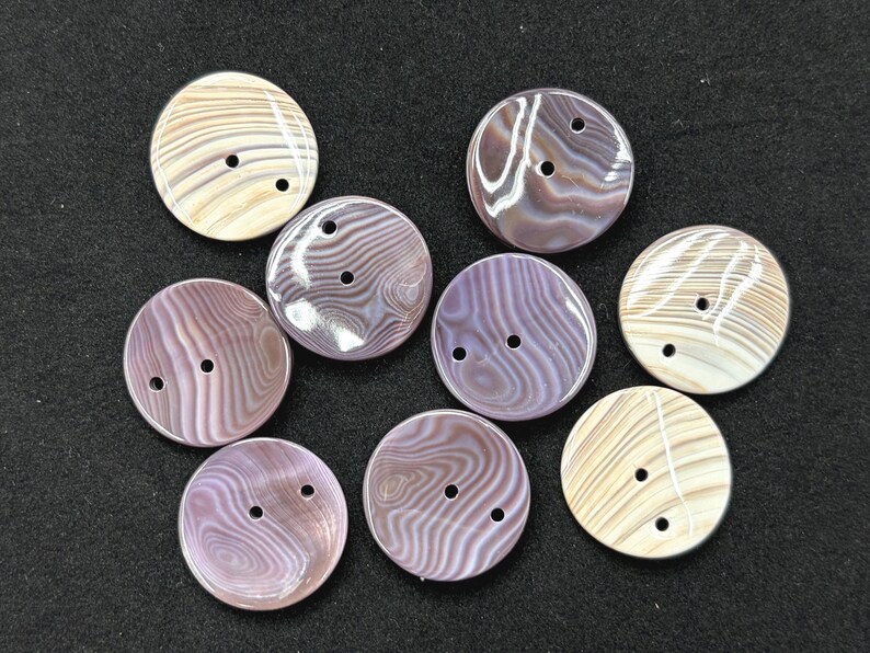 May include: A collection of eight round buttons featuring a smooth, glossy finish with swirling patterns in shades of beige, cream, and muted purple. Each button is circular and contains two small holes positioned centrally for attachment to fabric.