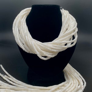 Peut inclure: Un collier de perles blanches en coquillage, disposé en forme de collier sur un mannequin noir. Les perles sont cylindriques et ont une finition lisse et polie.