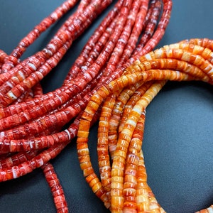 Puede incluir: Primer plano de dos hebras de cuentas de piedras preciosas rojas y naranjas. Las cuentas son redondas y tienen un acabado liso y pulido. Las cuentas están enhebradas en un cordón delgado y están dispuestas en un patrón suelto y fluido.