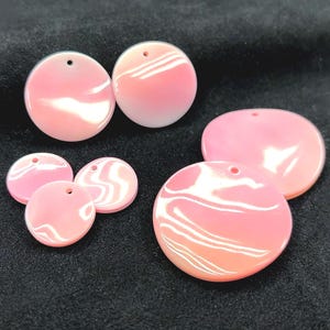 May include: A collection of six circular, smooth, pink shell pendants of varying sizes. Each pendant features a glossy, iridescent finish with subtle white swirling patterns and a single small hole at the top for threading or attachment purposes.