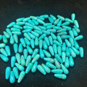 May include: A collection of turquoise teardrop-shaped beads. The beads are a vibrant turquoise colour with brown marbling. They are scattered on a dark surface, suggesting they are for jewellery making or crafts.