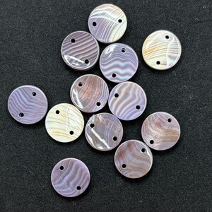 May include: A collection of twelve circular buttons featuring a smooth, iridescent surface with swirling patterns in shades of lavender, mauve, and cream. Each button has two small holes positioned near the top edge for attachment purposes.