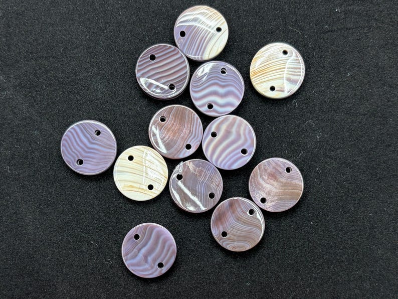 May include: A collection of twelve circular buttons featuring a smooth, iridescent surface with swirling patterns in shades of lavender, mauve, and cream. Each button has two small holes positioned near the top edge for attachment purposes.