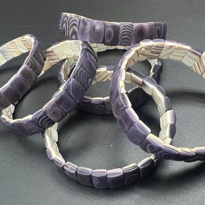 Op de afbeelding: Verschillende gestapelde armbanden met een uniek ontwerp. Elke armband bestaat uit kleine, rechthoekige kralen in tinten paars, wit en bruin. De kralen zijn in een cirkelvorm gerangschikt, wat een stijlvol accessoire creëert.