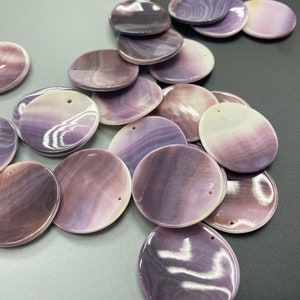 May include: A collection of circular, flat shell pendants featuring a polished, iridescent finish with swirling patterns in shades of deep purple, lavender, and white. Each pendant has a small, pre-drilled hole near the top edge for stringing.