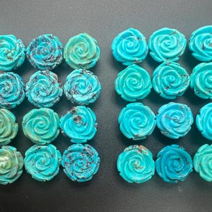 May include: Twenty-four turquoise-coloured rose-shaped gemstones. The carved stone roses vary in shades of blue and green, with some featuring dark veining. The gemstones are arranged in rows on a dark surface.