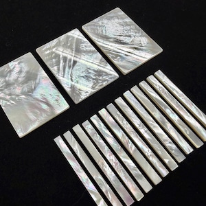 May include: A collection of rectangular and bar-shaped mother-of-pearl pieces. The rectangular pieces are larger, while the bar pieces are long and thin. The iridescent surfaces reflect light, showcasing a range of colors.