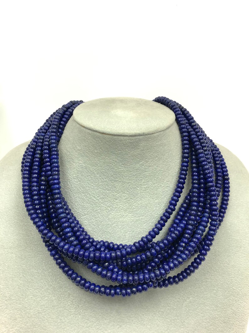 May include: A multi-strand necklace composed of small, round, deep blue beads. The necklace is displayed on a gray mannequin head, showcasing its layered design and rich color. The beads appear to be uniform in size and shape.
