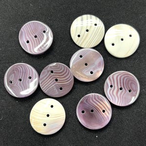 May include: A collection of nine round buttons featuring three holes each. The buttons display a variety of swirling patterns in shades of purple, mauve, and cream, with a smooth, polished finish across all pieces in the set.