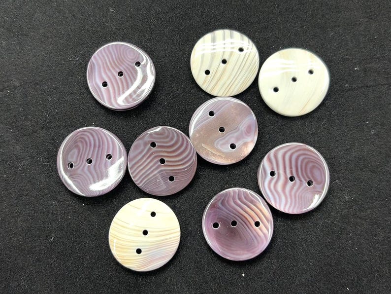 May include: A collection of nine round buttons featuring three holes each. The buttons display a variety of swirling patterns in shades of purple, mauve, and cream, with a smooth, polished finish across all pieces in the set.