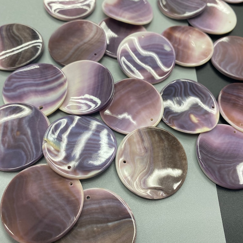 May include: A collection of circular, flat shell pendants featuring natural, swirling patterns in shades of purple, lavender, and white. Each smooth, polished disc has a single small hole drilled near the edge for hanging, showcasing a glossy, iridescent finish.