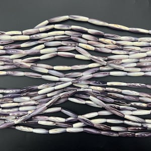 Pode incluir: Close-up de vários fios de contas alongadas em tons de roxo, branco e creme. As contas têm um acabamento brilhante e estão dispostas em um padrão em camadas sobre um fundo escuro. Estas contas são adequadas para fazer joias.