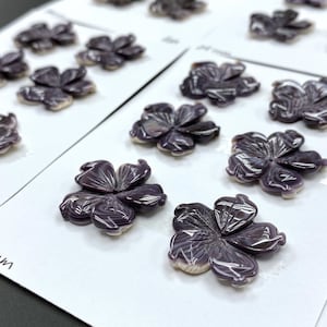 Puede incluir: Una colección de 10 adornos de flores de concha púrpura, cada uno con un diámetro de 25 mm. Las flores están dispuestas en cartulina blanca con texto negro que dice "25 mm".