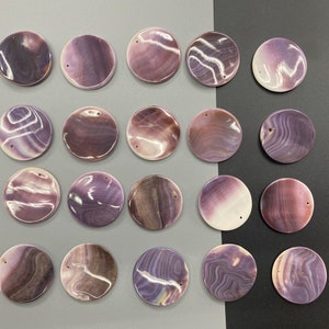 May include: A collection of twenty circular, flat shell pendants arranged in a grid. Each pendant features a unique, polished surface with swirling patterns in shades of purple, lavender, brown, and white. Each piece has a small drilled hole.