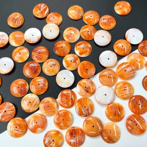 Op de afbeelding: Een verzameling ronde, oranje en witte schelpknopen met een natuurlijk, gestreept patroon. De knopen zijn ongeveer 2,5 cm in diameter.