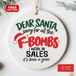 May include: A round white ceramic Christmas ornament featuring the text "Dear Santa, sorry for all the F-BOMBS I WORK IN SALES it's been a year." The design includes a red Santa hat icon, green and black lettering, and a gold hanging string.